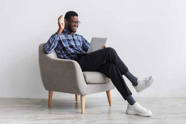 Молодий чорний чоловік, що має веб-дзвінок або відеоконференцію на ноутбуці, махає на камеру, сидить у кріслі на білій стіні
 - Фото, зображення