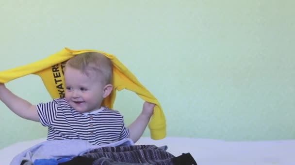 A cute baby is holding a huge stack of baby clothes and helping the mother with the housework. The boy is cleaning up the house. The child scatters things. Copy space- fun concept, organizing storage - Filmati, video