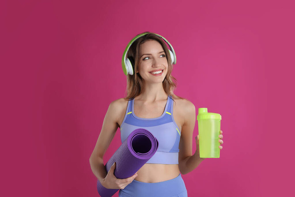Beautiful woman with headphones, yoga mat and shaker on pink background - 写真・画像