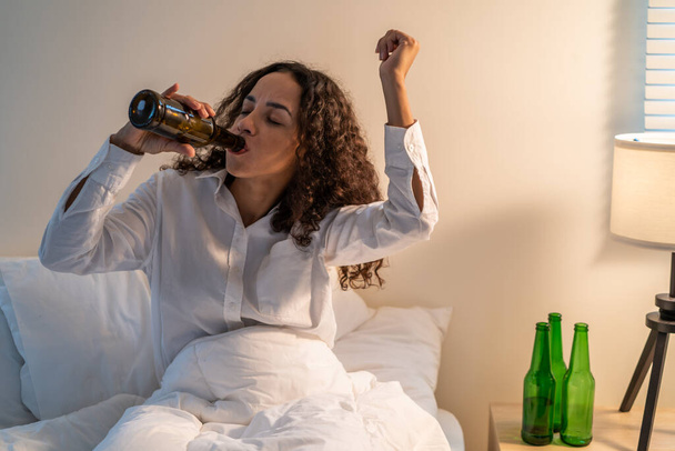 Drunk Latino woman hold beer bottle, feel hangover and dancing on bed. Young beautiful happy female stting in dark night in bedroom, feel alone and celebrate life problem with alcohol party at house. - Foto, immagini