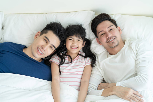 Portrait of handsome man gay family with young kid daughter in bedroom. Attractive romantic male lgbt couple lying down on bed with little adorable girl child in morning and looking at camera in house - Foto, Bild