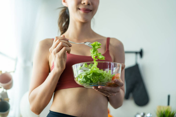Крупный план азиатской привлекательной женщины держать салат миску и есть овощи. Красивая спортивная девушка в спортивной одежде наслаждается чистыми овощами после физических упражнений для здоровья в доме. Концепция питания и здорового питания. - Фото, изображение