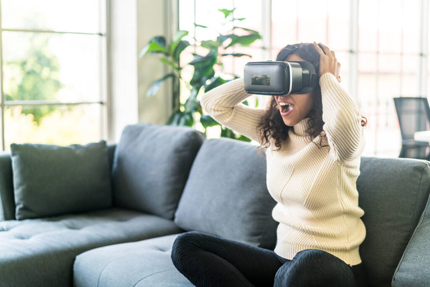 Latin woman using a virtual reality headset on sofa at home - Фото, изображение