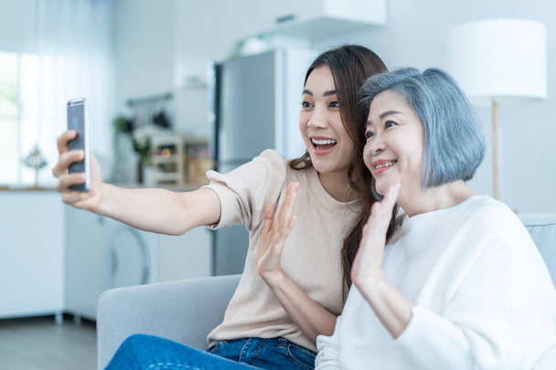 Asiatische glückliche Familie, ältere Mutter benutzt Mobiltelefon mit Tochter zu Hause. Attraktive schöne Mädchen sitzen mit älteren älteren Mutter auf dem Sofa und Videoanruf online kommunizieren Chat im Wohnzimmer im Haus. - Foto, Bild