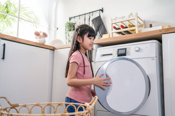 Asian young little kid put dirty clothes into washing machine in house. Lovely girl child sit on floor, feel happy to help family and loads laundry in washer appliance at home. Domestic-House keeping. - Zdjęcie, obraz