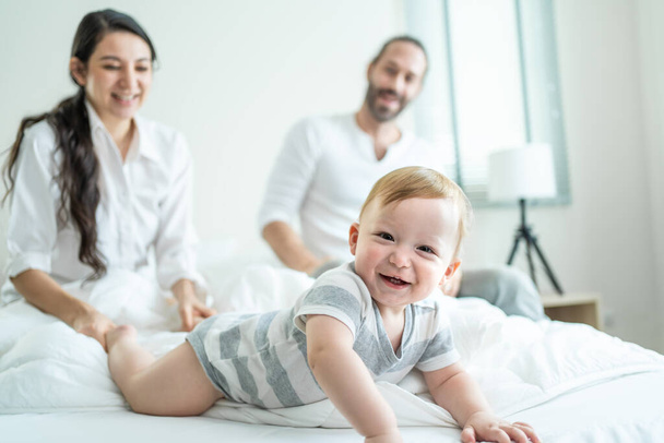 Caucasian parents play with cute baby boy child infant on bed at home. Happy family, attractive mom and dad look to toddler son crawl with happiness enjoy after wake up activity in bedroom in house. - Foto, imagen