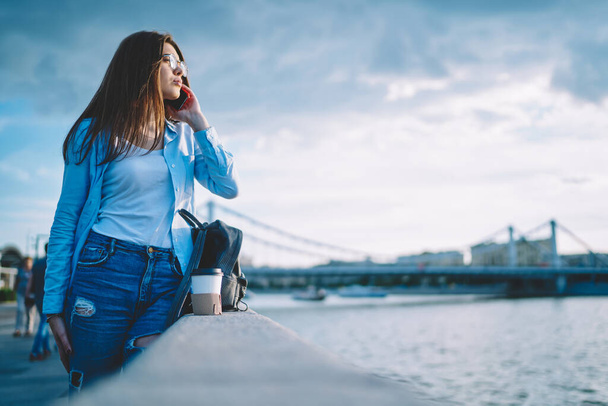 Attractive Caucaisan woman in spectacles using cellphone internet connection for communicate in roaming, millennial female tourist making international mobile conversation thinking at embankment - 写真・画像