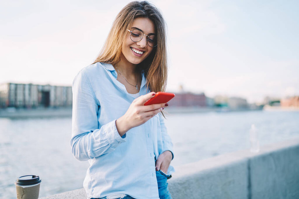 Young smiling woman in spectacles using smartphone for web networking during travel vacations, happy female tourist blogging using mobile phone and 4G internet mailing during sightseeing tour - Foto, Imagen