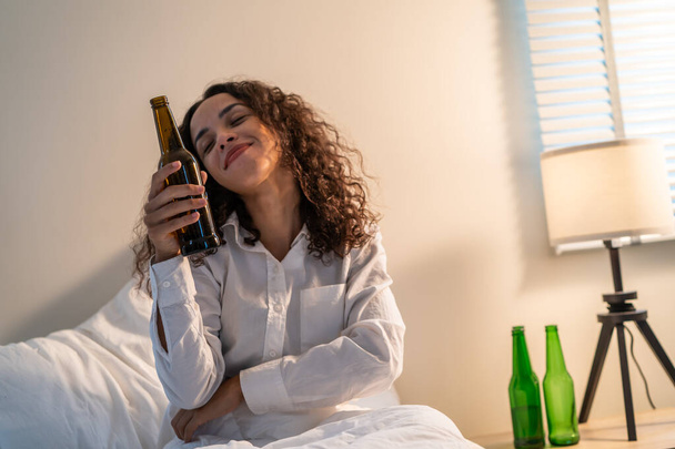 Betrunkene Latino-Frau hält Bierflasche in der Hand, verspürt Kater und tanzt auf dem Bett. Junge schöne glücklich weibliche Stechen in dunkler Nacht im Schlafzimmer, fühlen sich allein und feiern das Leben Problem mit Alkohol-Party im Haus. - Foto, Bild