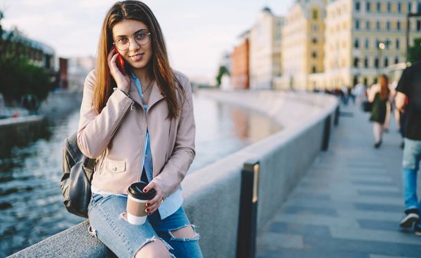 Portrait of attractive woman in trendy apparel making international mobile talk in roaming posing at embankment, millennial hipster girl with takeaway caffeine beverage using smartphone for calling - Photo, image