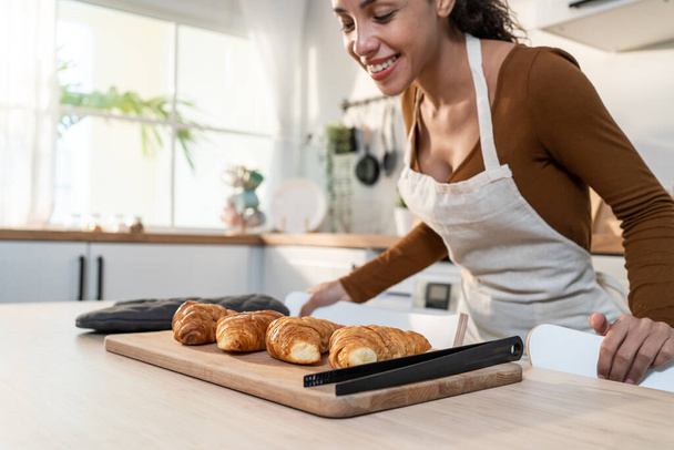 Aantrekkelijke jonge Latino vrouw bakken croissant op tafel in de keuken. Mooie vrouwelijke dragen schort gelukkig voelen en genieten van vrije tijd koken bakkerij thuis. Activiteit huisgemaakt in huis concept. - Foto, afbeelding