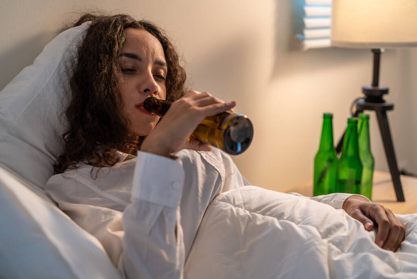 Opilá Latinoameričanka drží láhev od piva a pláče, cítí kocovinu na posteli. Mladé krásné deprese žena leží a pláče v slzách v ložnici, cítit srdce zlomené a oslavovat život s alkoholem v noci - Fotografie, Obrázek
