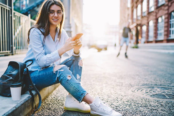 Portrait of brunette millennial student in stylish wear and eyewear posing in city during sunny day holding mobile phone, pretty Caucasian hipster girl with smartphone and backpack resting outdoors - Fotoğraf, Görsel