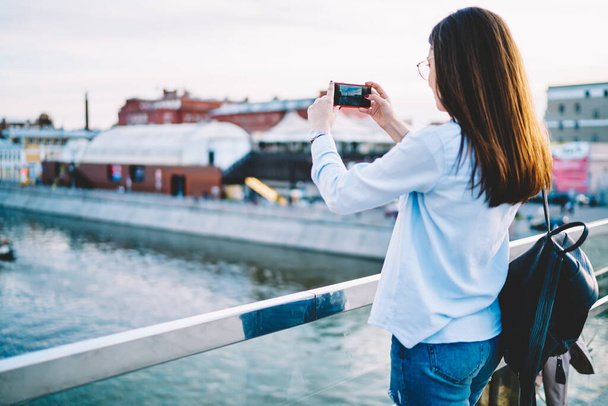 Обратный вид тысячелетней туристки, фокусирующей мобильную камеру на реке, делающей снимки на смартфоне, одетой в повседневную одежду девушки хипстера с рюкзаком, щёлкающей фото для обмена в блоге путешествий - Фото, изображение