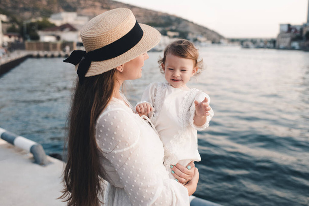 Feliz madre y la hija del bebé que se divierten usar vestido elegante blanco y sombrero de paja sobre la orilla del mar al aire libre. Mírense. Maternidad.  - Foto, imagen