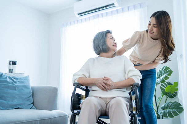 Asian young daughter support old disabled woman on wheelchair at home. Beautiful girl help and take care of senior elderly mature handicap mother patient doing physical therapy in living room in house - Photo, Image