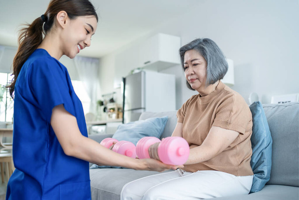 Asian Disabled old woman doing physiotherapist with support from nurse. Senior elderly handicapped female sit on sofa in living room using dumbbells workout exercise for patient in home nursing care. - Fotoğraf, Görsel