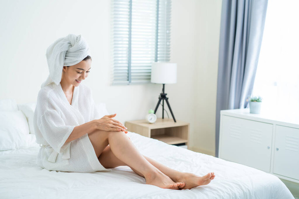 Asian beautiful woman put smooth and soft skin care on legs in bedroom. Attractive girl touches her knee and applying cream lotion for rejuvenation with gentle. Beauty, skin care and health concept. - Photo, Image