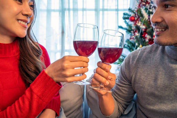 Asiático joven feliz pareja celebrar la fiesta de Navidad juntos en casa. Atractivo hermoso hombre y mujer bebiendo alcohol de vino tinto y vidrio tostado en el sofá, siéntase feliz y emocionado por la Navidad en casa. - Foto, imagen