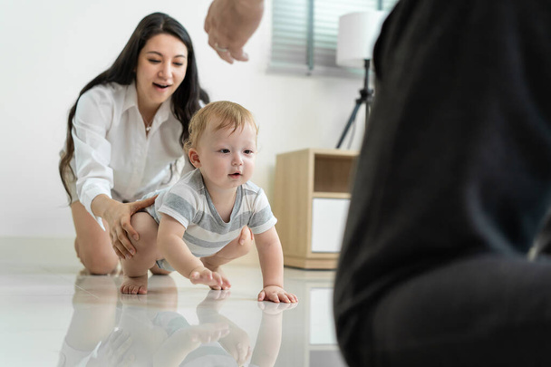 Caucasian baby boy child crawling with parents support in house. Happy family, mother and father help young toddler son learn how to walk and take step on floor to develop skill in living room at home - Foto, Bild