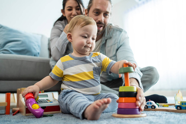 Caucasian loving parent looking at baby toddler playing in living room. Attractive couple mother and father watch young little infant son child's development. Activity relationship at home in house. - Photo, image