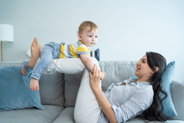 Caucasian loving mom play with baby boy child on sofa in living room. Happy family, attractive beautiful caring young mother lifting toddler son fly up enjoy doing airplane game in morning in house. - Foto, imagen