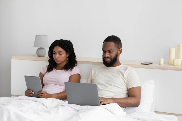 Busy serious millennial black wife and husband working on laptop and tablet, sitting on bed - Фото, изображение