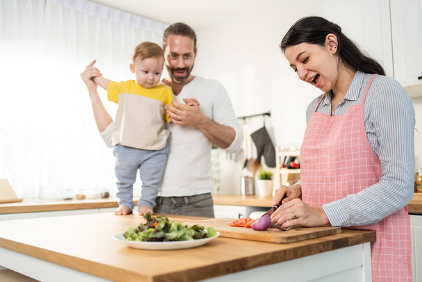 Caucasian beautiful parents cook food with baby boy toddler in kitchen. Happy family, Attractive young mother making healthy salad for lunch while husband father holding little kid son infant in house - Foto, Imagen