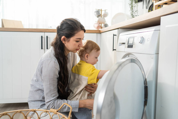 Caucasian young mom doing housework with baby boy toddler in kitchen. Beautiful mother doing laundry, put clothes to washing machine while take care of her son little kid in house. Family-housekeeping - Фото, изображение