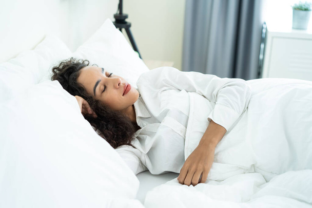 Latino beautiful woman lying down on bed in bedroom in early morning. Attractive young female in pajamas sleeping alone on comfortable pillow and cozy blanket in bright room for health care at home. - Photo, Image