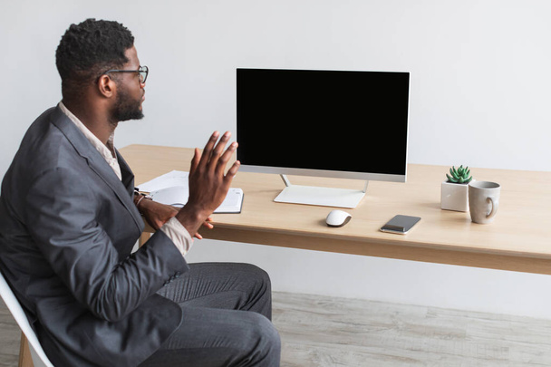 Young black entrepreneur having online business meeting at office, waving at computer monitor with empty screen, mockup - Фото, изображение