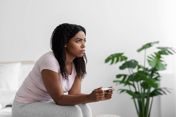 Απογοητευμένη νεαρή μαύρη γυναίκα κάθεται στο κρεβάτι και κοιτάζοντας αρνητικό τεστ εγκυμοσύνης έχουν προβλήματα υγείας των γυναικών - Φωτογραφία, εικόνα