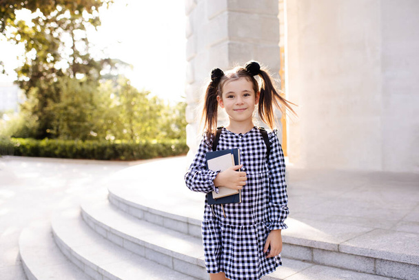 Sweet smiling young child girl 5-6 year old wear checkered black and white dress and backpack holding books stand outdoors close up. Looking at camera. Back to school. Study concept.  - Фото, зображення