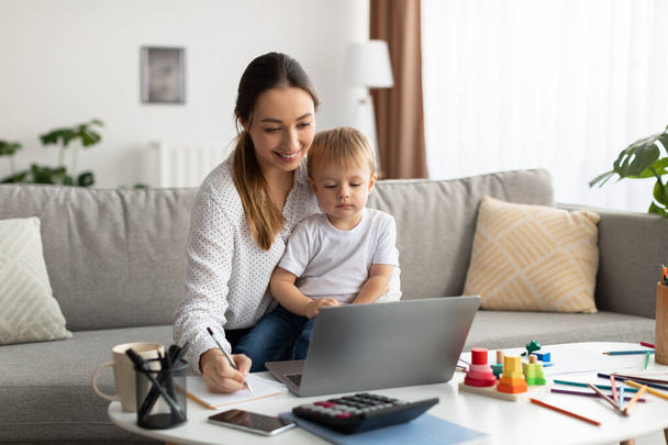 Online education. Young mother holding baby and studying with laptop, taking notes to notepad while watching webinar - Foto, Bild