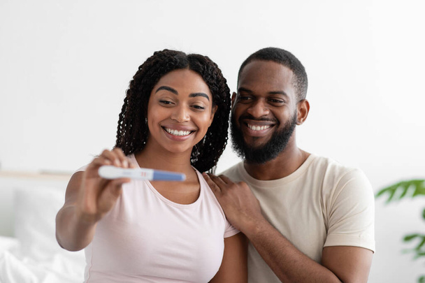 Satisfied attractive millennial black woman and guy show positive pregnancy test at camera in bedroom interior - Fotó, kép