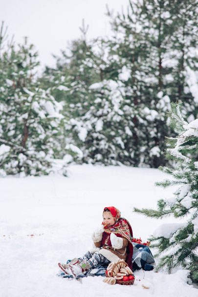 Beautiful little child girl plays in snowy forest near basket with apples and bagels. She dressed in the old Russian style in red headscarf. - Valokuva, kuva