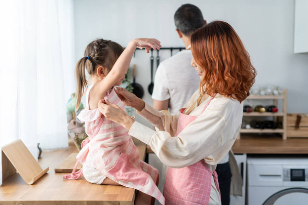 Asian beautiful young mother cooking foods with baby kid in kitchen. Happy family spend time on holiday together in house, loving parent tying apron for little child daughter prepare to make breakfast - Foto, afbeelding