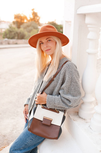 Stylish blonde young adult girl 22-24 year old wear felt hat, denim pants and knit woolen cardigan holding trendy leather bag walk in city street posing over sunset background outdoors close up. 20s. - Foto, Bild