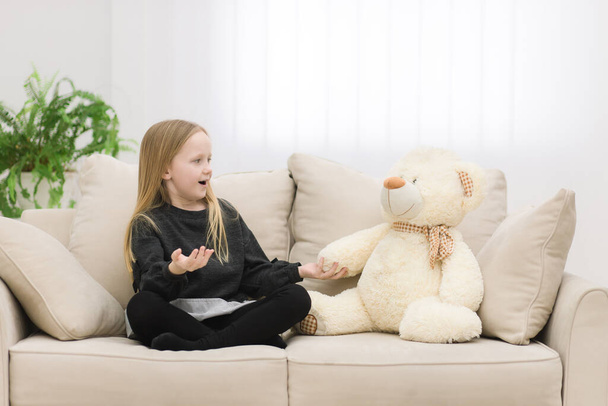 Photo of teddy bear and blonde girl looking on each other on the sofa. - Zdjęcie, obraz
