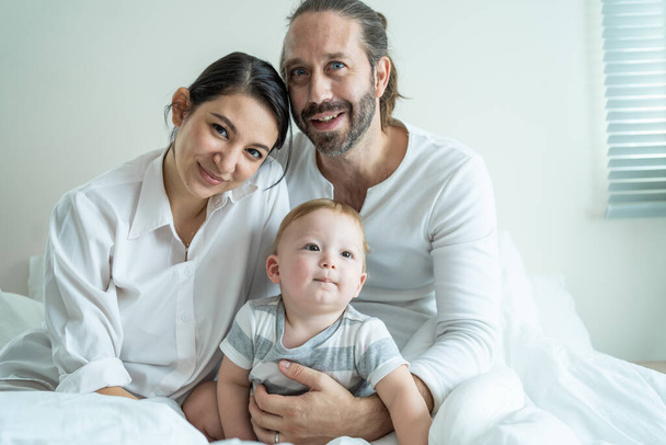 Портрет кавказской счастливой семьи, улыбающейся, смотрите в камеру в спальне. Молодые привлекательные родители, отец и мать сидят на кровати с маленьким мальчиком и наслаждаются утренним бодрствованием дома. - Фото, изображение