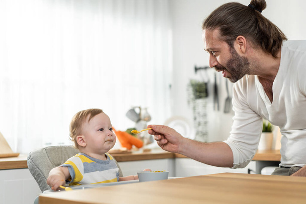 Caucasian beautiful father take care of baby boy toddler in kitchen. Happy family, Attractive loving daddy cook and feeding healthy foods to little kid son infant while sit on chair for lunch in house - Фото, зображення