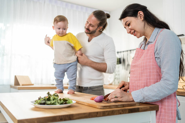 Caucasian beautiful parents cook food with baby boy toddler in kitchen. Happy family, Attractive young mother making healthy salad for lunch while husband father holding little kid son infant in house - Foto, Bild