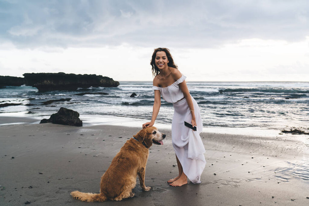 Full length of excited barefoot female in white clothes smiling and looking at camera while walking with dog on beach - Foto, Imagem