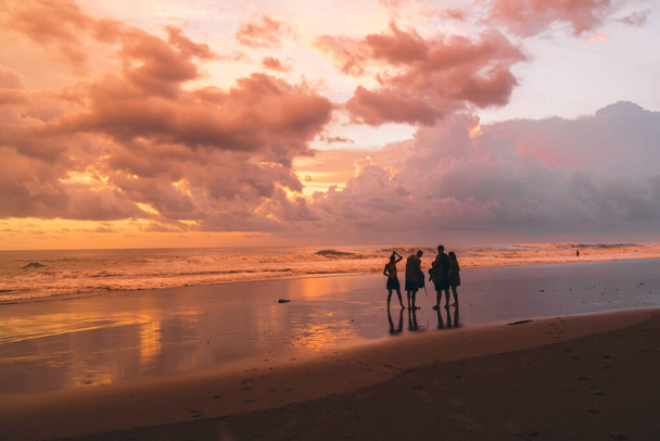 Back view of unrecognizable tourists standing on wet sandy shore and talking with each other while enjoying amazing orange sunset - Foto, imagen