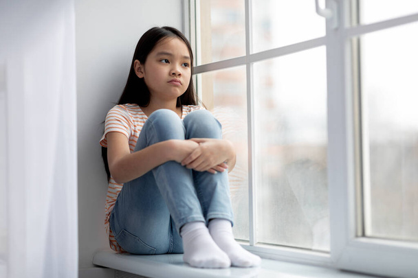 verveeld aziatisch meisje zitten op vensterbank alleen, kopiëren ruimte - Foto, afbeelding