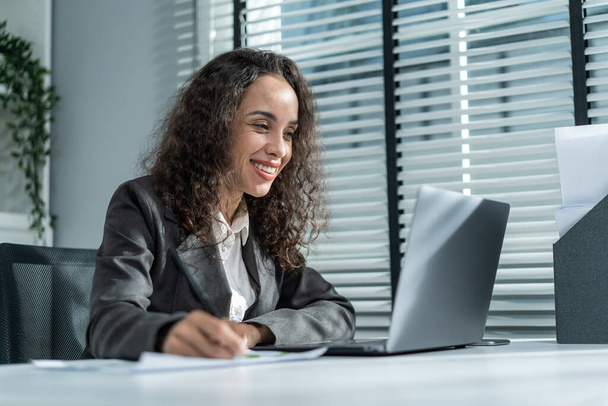 Happy beautiful Latino businesswoman in formal suit work in workplace. Attractive female employee office worker smile, use laptop computer for discuss with co worker and take note on paper in company. - Fotografie, Obrázek