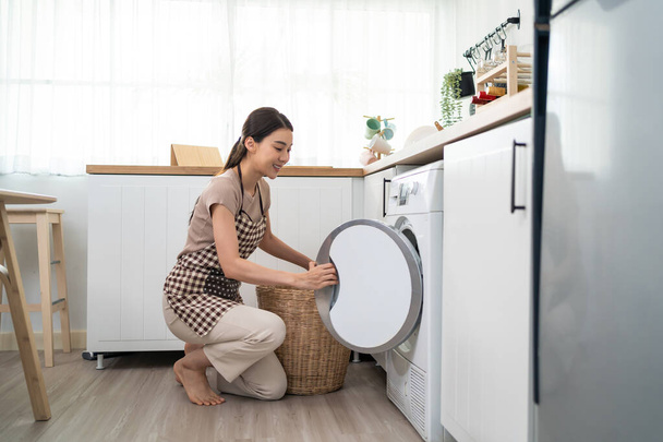 Asian beautiful woman put dirty clothes to washing machine in house. Attractive girl wear apron sit on floor, feel happy to loads laundry in washer appliance at home. Domestic-House keeping concept. - Fotoğraf, Görsel
