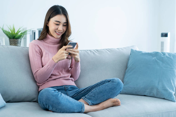 Asian beautiful woman sit on sofa and chat on mobile phone in house. Happy attractive young girl spend leisure time at home, feel relax and enjoy communicate and discuss on smartphone in living room. - Valokuva, kuva