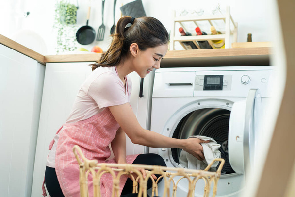 Asiatische schöne Frau legte schmutzige Wäsche in die Waschmaschine im Haus. Attraktive Mädchen in Schürze sitzen auf dem Boden, freuen sich, Wäsche in die Waschmaschine zu Hause zu laden. Häusliches Pflegekonzept. - Foto, Bild