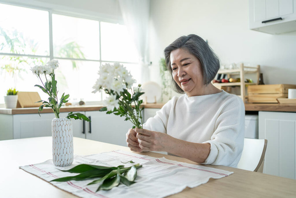 Asian senior old woman puts flowers on vase with happiness in kitchen. Beautiful active elderly grandmother sit on table, feeling happy while decorate and renovate apartment. Activity at home concept. - Foto, Bild